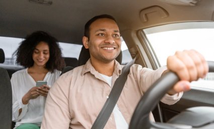 A driver is focused on the road while a passenger in the back seat uses a smartphone. Both faces are blurred.